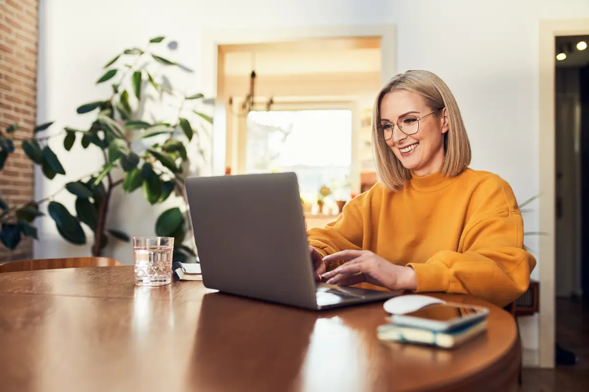 A smiling woman in an orange sweater and glasses works on a laptop at a wooden table.