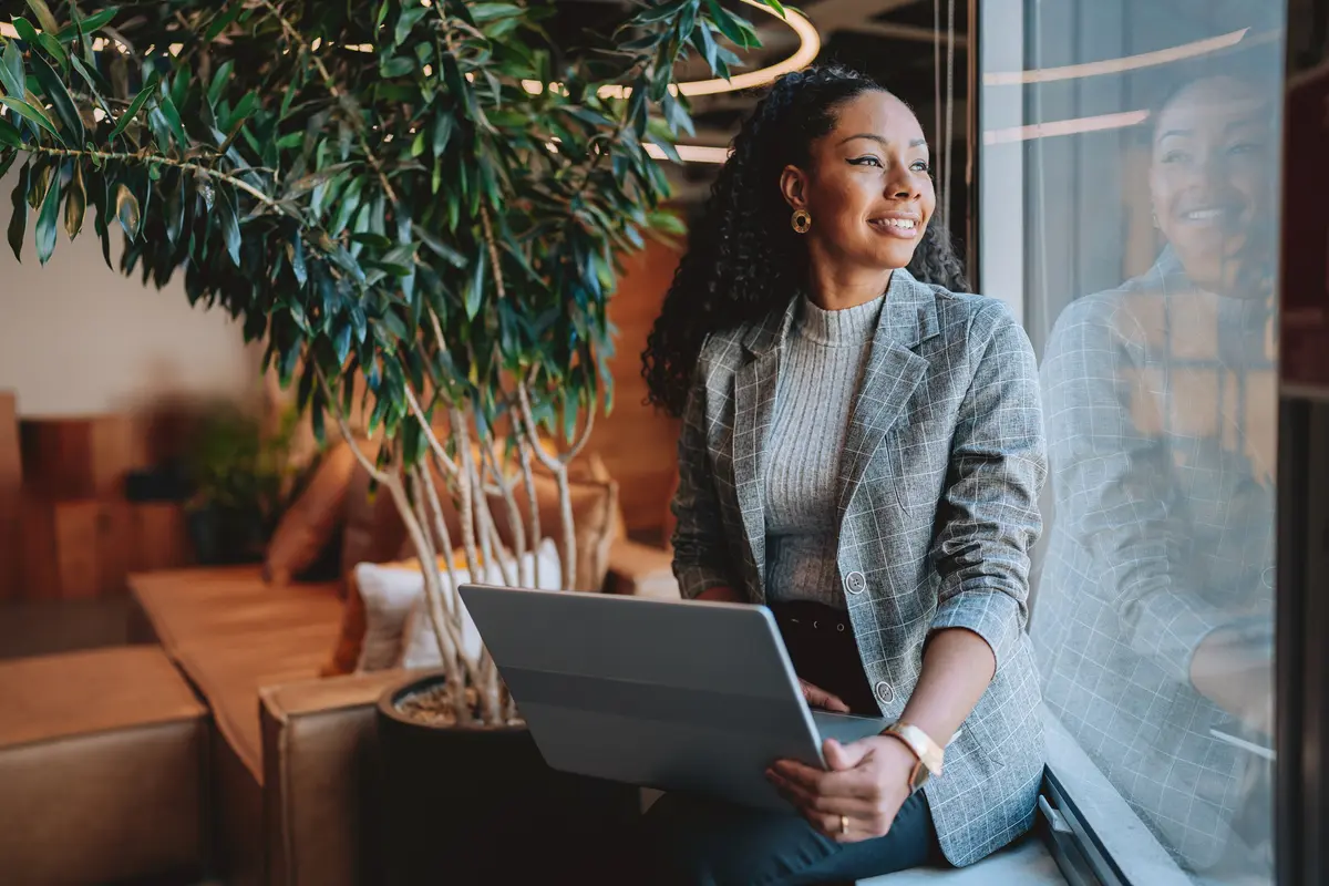 Smiling woman in a gray blazer holds a laptop while gazing out a window near an indoor plant.