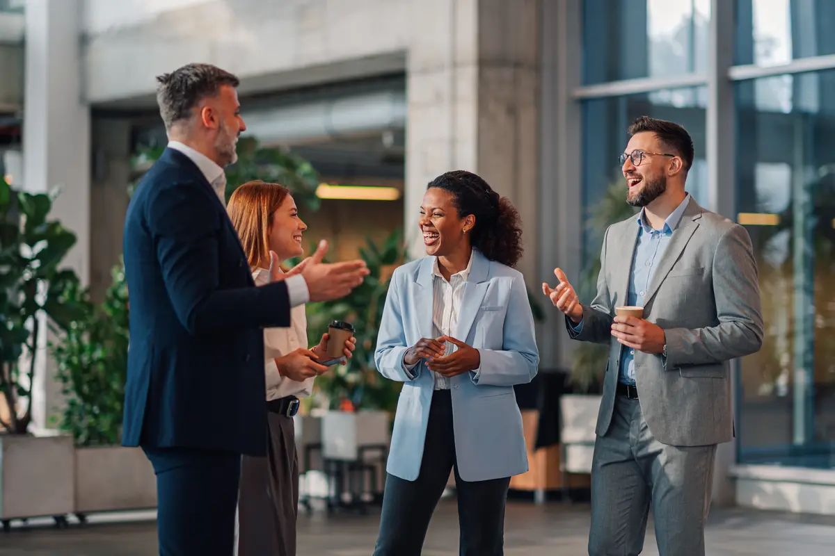 Four business professionals in suits laugh together while holding coffee cups in a modern office lobby.