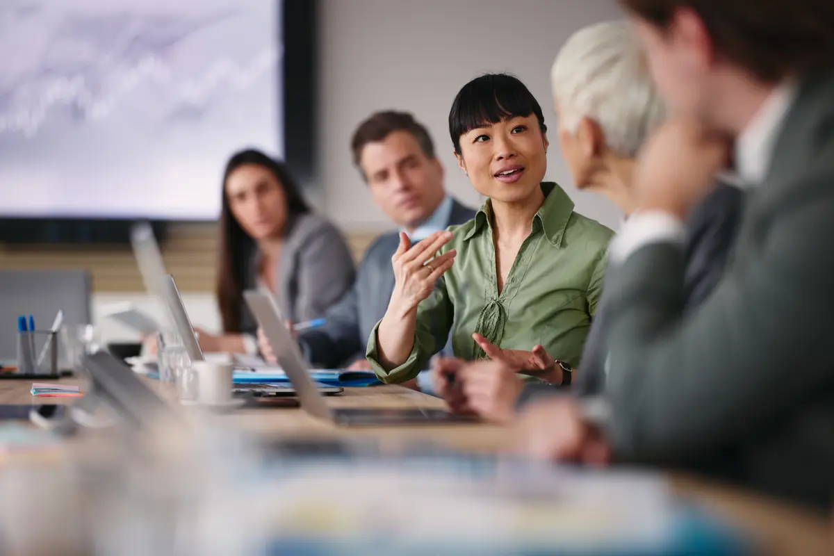 A woman in a green blouse gestures while speaking to colleagues around a conference table during a meeting.