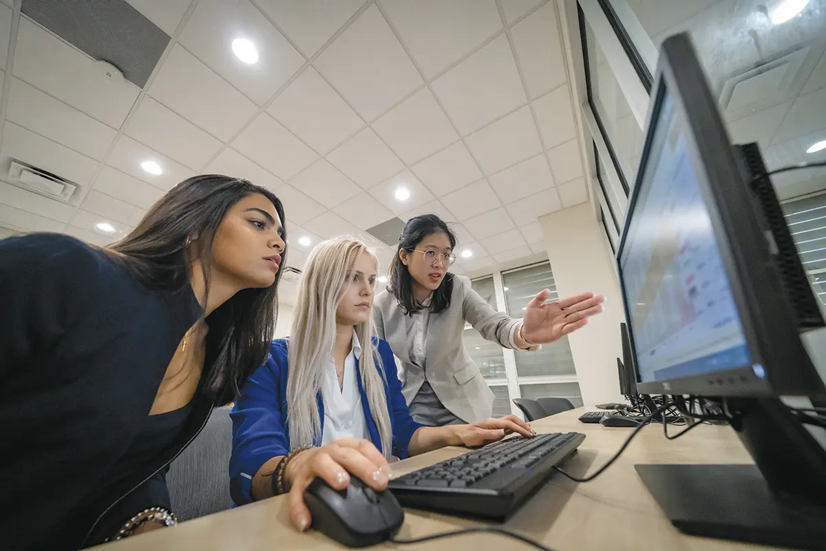 Three young women collaborate at a computer workstation, one pointing at the screen while others watch attentively.