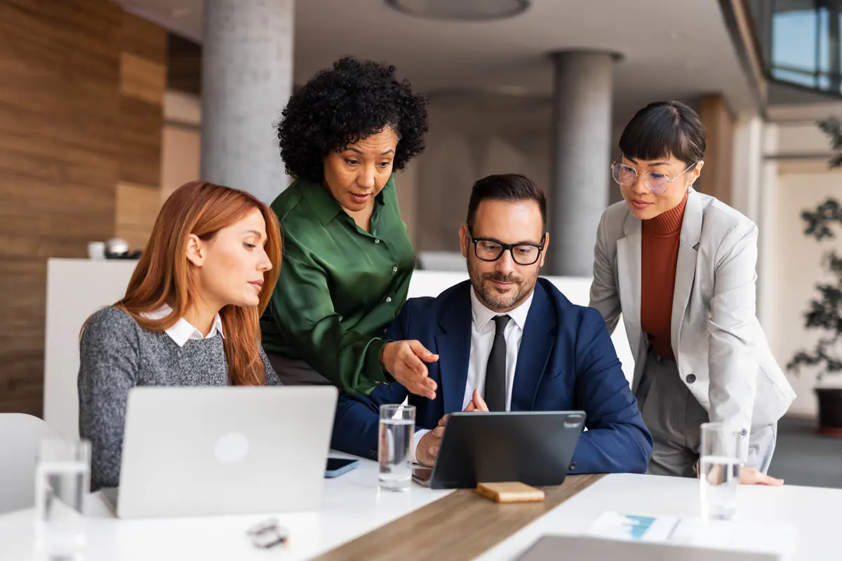 Four business professionals gather around a tablet and laptop, collaborating during a meeting in a modern office.
