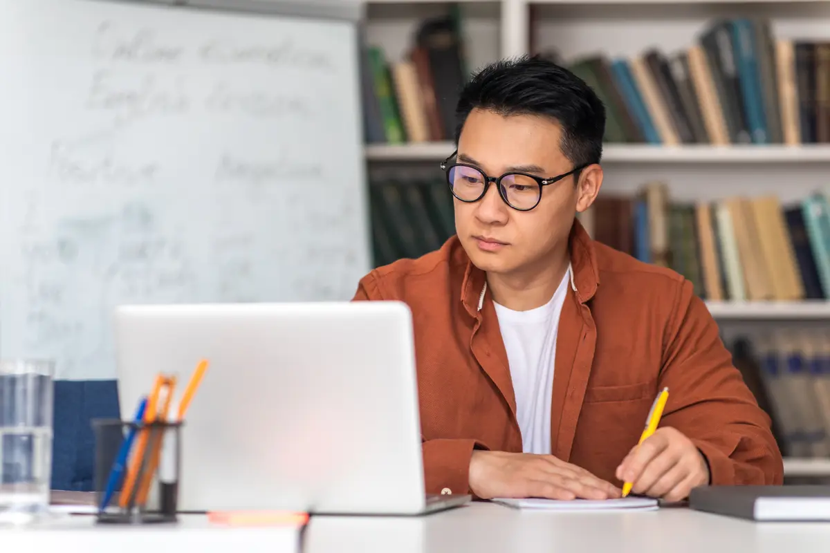 A man in glasses takes notes while looking at a laptop in a library setting.