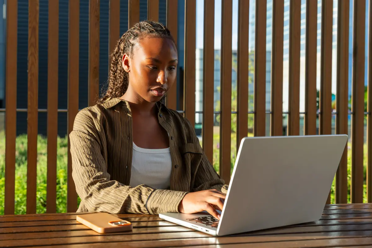 A young woman in an olive shirt works on a laptop at an outdoor wooden table in sunlight.