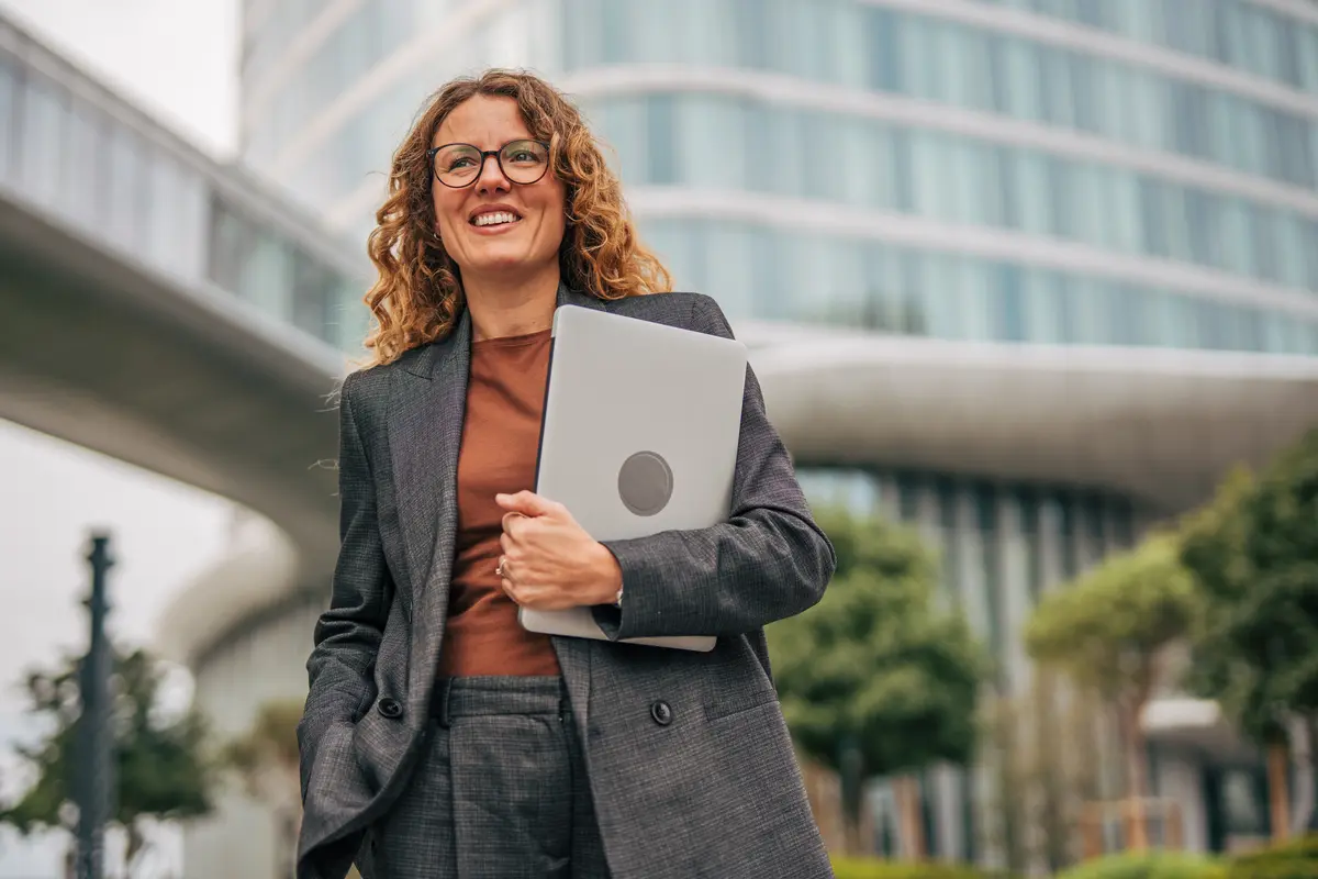 Smiling woman in gray suit holding a laptop while standing outside a modern glass office building